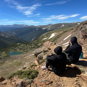 young adults standing at a scenic overlook in the American West with a backpack, mountains in the background — representing the work and travel lifestyle offered by AdventureEXP.
